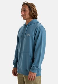 Blue hoodie made of soft fabric, featuring a front pocket, drawstring hood, and small logo on the chest. Long sleeves with ribbed cuffs.