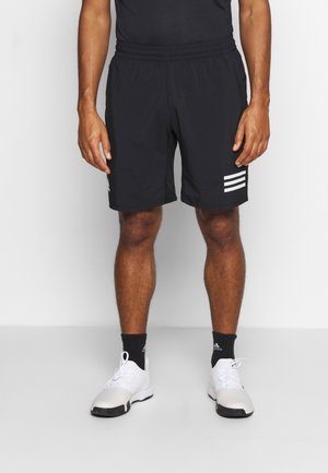 Homme portant un short de sport noir avec un logo blanc à trois bandes, des chaussettes noires Adidas et des chaussures de sport blanches, debout.