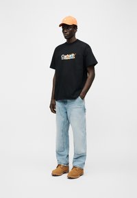 Άνδρας που φορά πορτοκαλί καπέλο, μαύρο T-shirt με σχέδιο, ανοιχτόχρωμα τζιν με μπαλώματα και καφέ μπότες εργασίας, στέκεται μπροστά σε λευκό απλό φόντο.