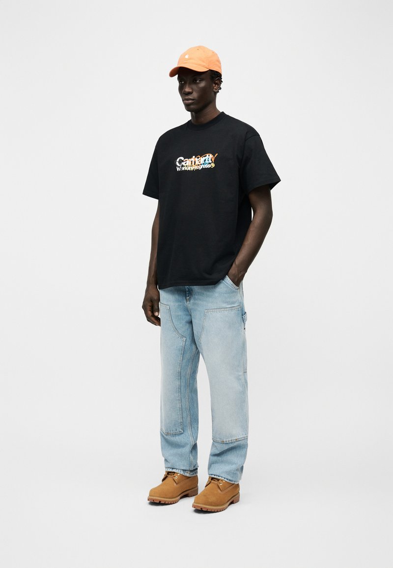 Homme portant une casquette orange, un t-shirt noir à motif, un jean patchwork bleu clair, et des bottes de travail couleur tan, debout devant un fond blanc uni.