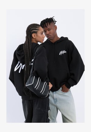 Le felpe nere oversize presentano un logo bianco sulla parte anteriore e posteriore. Il modello tiene una borsa nera con strisce bianche, abbinata a jeans strappati.