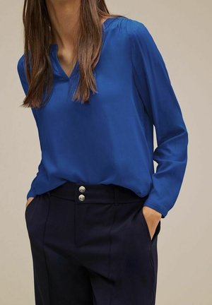 Blouse bleue à manches longues avec un col en V ; associée à un pantalon bleu foncé avec deux boutons à la taille. Texture douce.