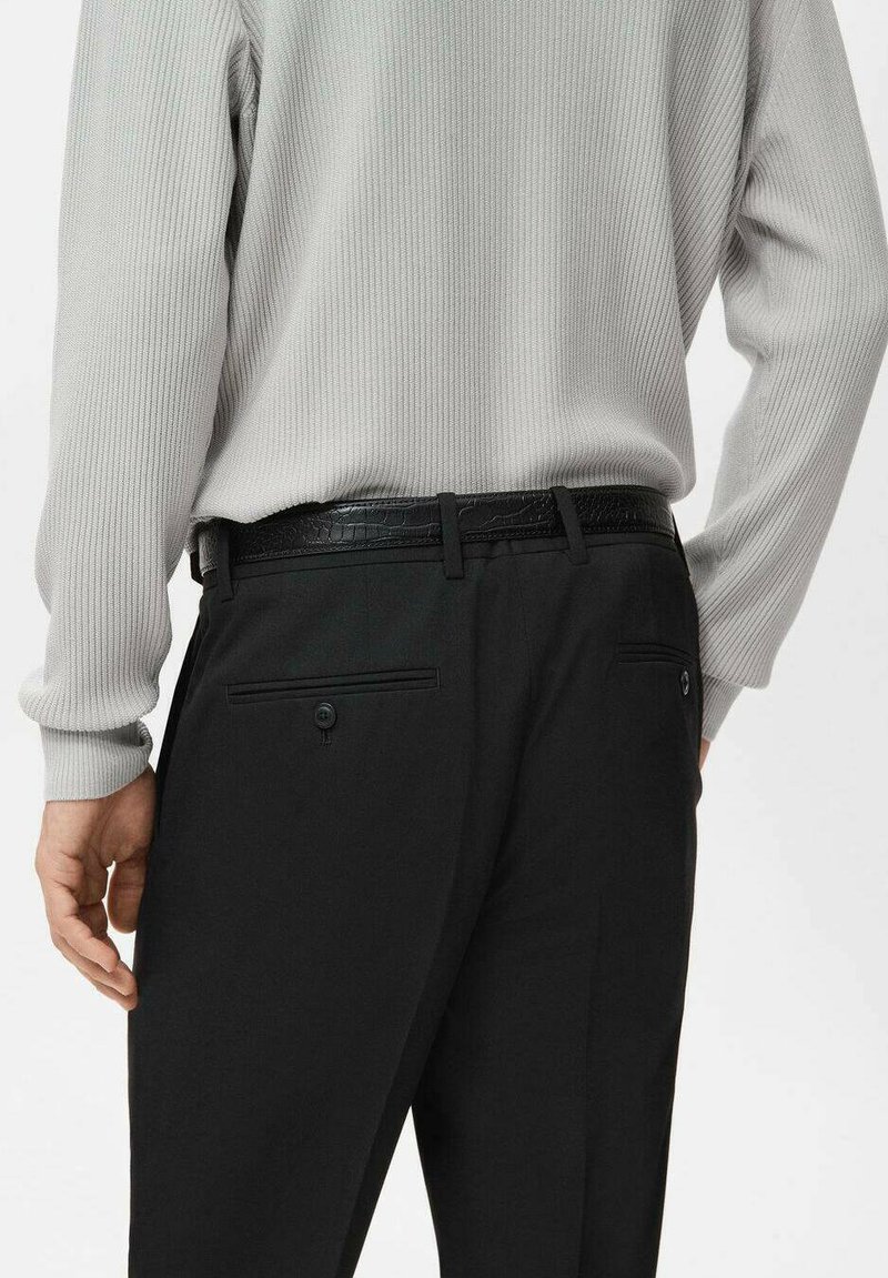 Mann trägt einen hellgrauen, gerippten Pullover, der in schwarze Anzughose mit einem schwarzen, strukturierten Gürtel gesteckt ist, gezeigt von hinten ab der Taille abwärts.