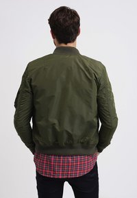 Maslinasto zelena bomber jakna s rebrastim rukavima i donjim rubom, zatvorenim bočnim džepovima i glatkim, laganim materijalom. Nosena preko crvene karirane košulje.