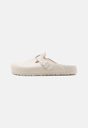 Biały chodak typu slip-on z regulowanym paskiem na klamrę i teksturowaną podeszwą, zaprojektowany do codziennego użytku.