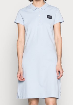 Frau trägt ein hellblaues Kurzarm-Polokleid mit Frontknöpfen und einem Tommy Hilfiger Logo-Patch auf der Brust.