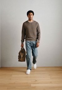 Πλεκτό πουλόβερ με καφέ και μπεζ ρίγες, μπλε τζιν και λευκά αθλητικά παπούτσια· κρατώντας καφέ τσάντα· στέκεται πάνω σε ξύλινο πάτωμα μπροστά σε απλό φόντο.