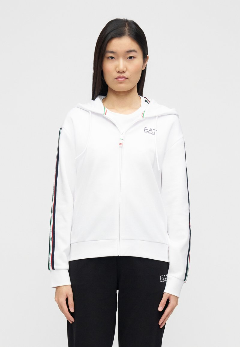 Λευκή φούτερ ζακέτα με φερμουάρ, κατασκευασμένη από μαλακό ύφασμα, με κουκούλα και πολύχρωμες ρίγες στα μανίκια. Συνδυάζεται με μαύρο παντελόνι.