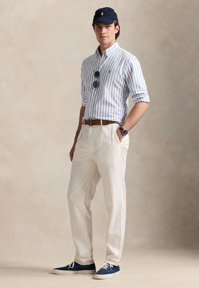 Bavlněná pruhovaná košile na knoflíky v světle modré a bílé, béžové kalhoty, námořnická čepice, modré tenisky a sluneční brýle. Hnědý pásek s hodinkami.