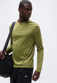 Άνδρας που φοράει πουκάμισο μακρυμάνικο ελιάς και μαύρο παντελόνι, κρατώντας μαύρη τσάντα ώμου, κοιτά προς τα αριστερά του με φόντο απλό.