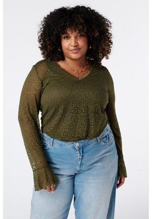 Femme aux cheveux bouclés portant un chemisier transparent à motif vert olive et un jean bleu clair, debout devant un fond blanc uni.