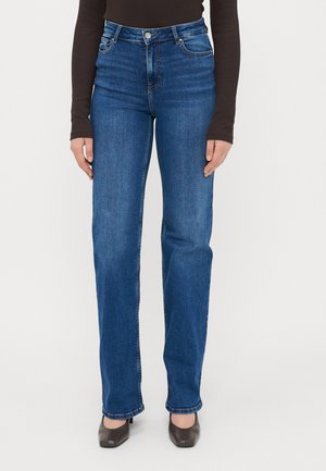 Jeans de ganga azuis de cintura alta com pernas retas, apresentando cinco bolsos e detalhes de costura subtil. Combinados com sapatos castanhos escuros.