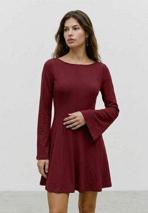 Femme portant une robe bordeaux à manches longues avec des poignets évasés, debout sur un fond clair uni, regardant légèrement de côté.