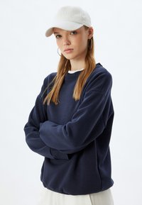 Sudadera amplia de color navy con puños y cuello acanalados, combinada con una gorra de béisbol blanca. La modelo tiene el cabello largo y lleva pendientes de aro.