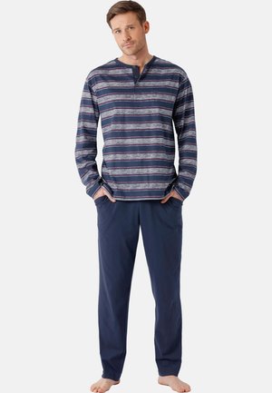 Mann steht barfuß, trägt dunkelblaue Hose und ein langärmliges, gestreiftes Hemd mit Knöpfen, die Hände in den Taschen, vor einem schlichten Hintergrund.