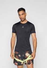 Sort kortærmet sportstrøje med rund halsudskæring og lille hvidt logo, parret med multicolorer shorts med abstrakte mønstre og gule accenter.