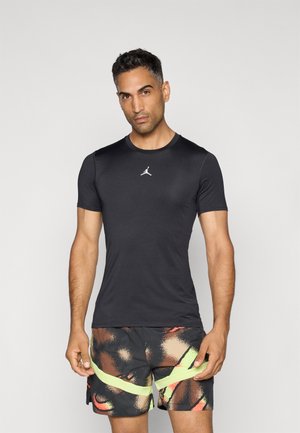 Schwarzes kurzärmliges Sportshirt mit rundem Ausschnitt und kleinem weißen Logo, kombiniert mit mehrfarbigen Shorts, die abstrakte Muster und gelbe Akzente aufweisen.