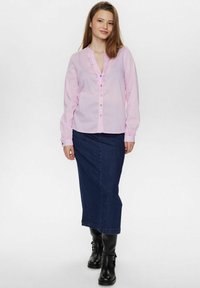 Camicia button-up rosa chiaro con colletto arricciato e bottoni colorati, abbinata a una lunga gonna di jeans scura e stivali neri alla caviglia.