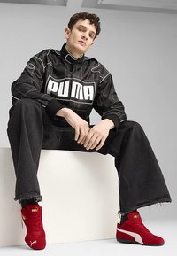 Must musta jakk valge "PUMA" logoga, laiad mustad teksad narmendava äärega ning punased seemisnahast tennised valgete detailidega. Modell istub helel pinnal.