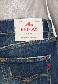Vaqueros de mezclilla azules con una etiqueta blanca que lleva un texto rojo que dice "REPLAY EST. 1981", costuras visibles y una textura desgastada en la tela.
