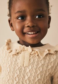 Smiling young child wearing a cream cable-knit sweater with ruffled collar and velvet bow, looking off-camera against a neutral background.