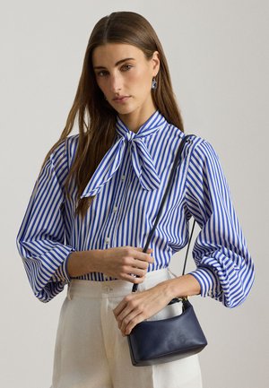 Γυναίκα που φοράει μπλούζα με μπλε και λευκές ρίγες και γραβάτα, λευκό παντελόνι, κρατώντας μια μικρή μαύρη δερμάτινη τσάντα στον ώμο της.