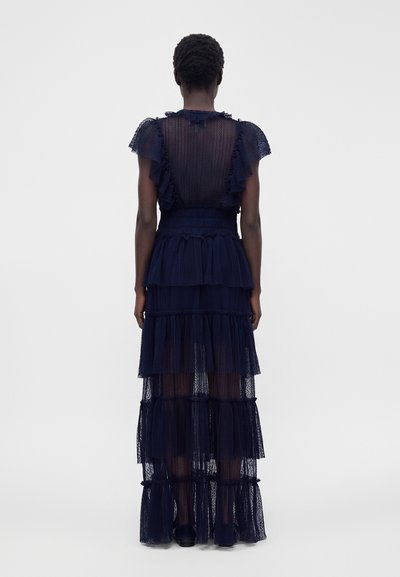 Femme aux cheveux courts portant une robe en dentelle transparente bleu marine à étages avec des manches courtes à volants, debout de dos sur un fond uni.