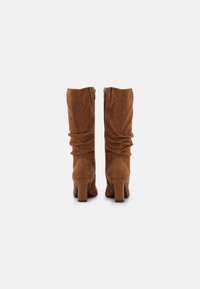 Brązowe zamszowe kozaki za kolano z marszczonym wzorem, solidnymi obcasami typu blok i zamkami z tyłu. Teksturowane wykończenie podkreśla atrakcyjność wizualną.