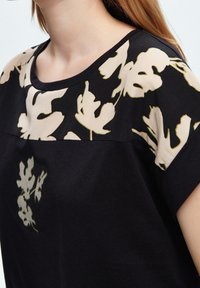 Svart topp med korta ärmar, med en beige blommösdesign vid halsringningen och bröstområdet. Slätt tyg med en avslappnad passform.