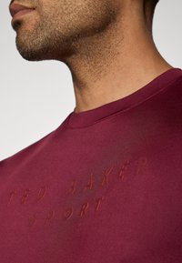 Bordo medvilnės marškinėliai su apvalia kaklo linija, su įspauditu tamsesniu "TED BAKER SPORT" tekstu. Lygi tekstūra su minimaliu blizgesiu.