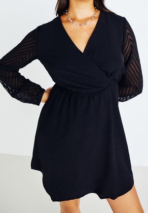 Black V-neck dress with sheer long sleeves featuring zigzag patterns, elastic waist, worn with a silver chain necklace.
