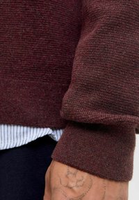 Burgundowy sweter z dzianiny o fakturowym wykończeniu, zrolowanym mankiecie i warstwowym pod spodem podkoszulkiem w paski. Widoczna ręka i tatuaż.
