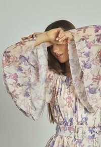 Blumenkleid mit transparenten, glockenförmigen Ärmeln in Pastellfarben. Es hat einen gerafften Ausschnitt und einen passenden Gürtel mit einer quadratischen Schnalle.