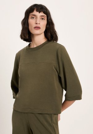 Woman with shoulder-length dark hair wearing an olive green loose-fit top with elbow-length sleeves and matching pants standing against a plain background.