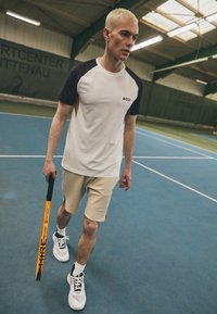 Bílé sportovní tričko s černými rukávy, béžové šortky, drží oranžovou pálku, na modrém tenisovém kurtu se zeleným pozadím.