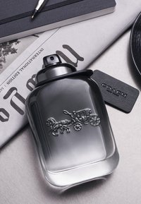 Bouteille de parfum en verre transparent avec un dégradé sombre, présentant un motif en relief de cheval et de carrosse ; elle inclut une étiquette noire portant l'inscription "COACH."