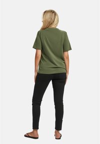 Woman with long blonde hair wearing an olive green short-sleeve shirt, black slim pants, and black sandals, standing facing away.