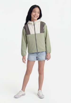 Smilende pige, stående, iført en grøn og beige hættetrøje med lynlås, lyse denimshorts og hvide sneakers mod en ensfarvet hvid baggrund.