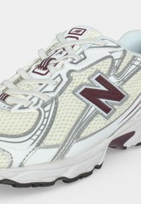 Близък план на бяла и сива маратонка New Balance с бордо лого и акценти и текстилна горна част с шнурове.