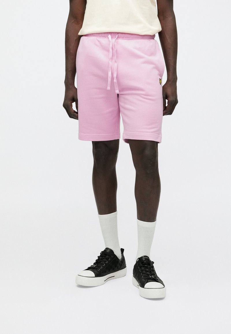 Rózsaszín pamut rövidnadrág elasztikus derékrésszel és húzózsinórral, oldalzsebekkel és egy kis fekete logóval a bal lábszáron. Fekete tornacipő és fehér zokni.