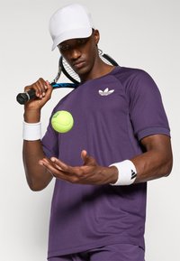 Violetti urheilupaita, jossa on valkoinen Adidas-logo, valkoiset käsivarsinauhat ja valkoinen lippis. Pitää kädessään tennismailaa, jonka yllä leijuu keltainen tennispallo.