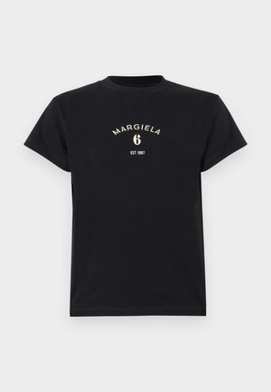 Femme tatouée portant un t-shirt noir Margiela, un pantalon large noir, des bottes noires et tenant un petit sac à main noir.