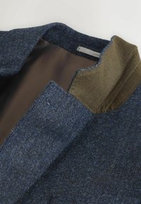 Wollblazer mit einem blauen und grauen strukturierten Gewebe, einem kontrastierenden olivgrünen Kragen und einem glatten braunen Innenfutter.