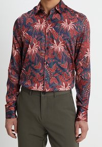 Chemise à manches longues avec un fond bleu foncé orné de motifs de feuilles tropicales rouges et blanches, fermeture boutonnée à l'avant et col classique.