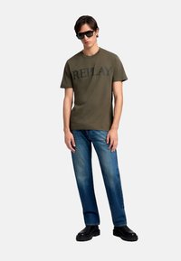 Olivengrøn t-shirt med "REPLAY" trykt i mørk skrift, parret med lyseblå jeans og sorte chunky sko. Modellen bærer solbriller.