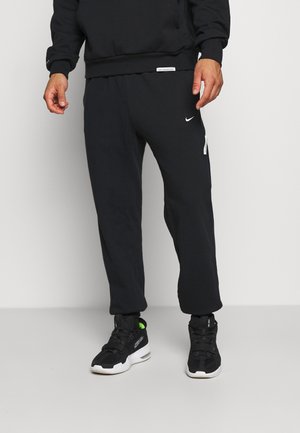Mężczyzna ubrany w czarne spodnie dresowe Nike i czarne buty sportowe z białymi podeszwami, stojący na tle jednolitego, jasnego tła.