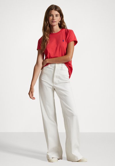 T-shirt di cotone rosso con maniche corte e un logo blu sul petto, abbinato a pantaloni bianchi a vita alta e gamba larga e scarpe color crema.