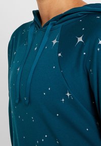 Hoodie teal avec cordon de serrage, orné de motifs d'étoiles argentées. Fabriqué en tissu doux avec une texture lisse et une coupe décontractée.