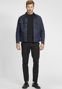Temno modra denim jakna z dvema žepek na prsih, nošena čez črno srajco, kombinirana z črnimi hlačami in sijočimi črnimi čevlji.