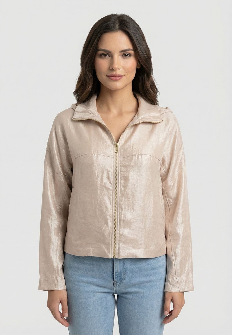 Femme aux cheveux foncés portant une veste beige brillante à fermeture éclair et un jean bleu clair, debout devant un fond blanc uni.
